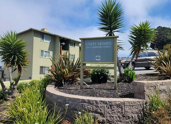 A sign for "UNITY HOMES" stands in front of a building with a landscaped area.