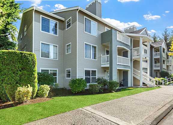 Gray apartment building with balconies, walkways, and lush green landscaping.