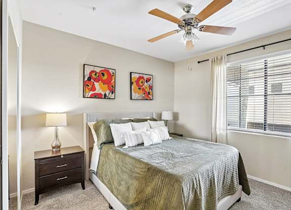 A bedroom with a bed, two lamps, a ceiling fan, and two paintings on the wall.