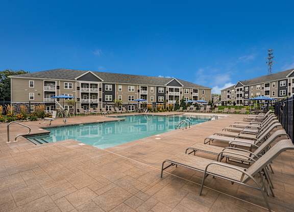A swimming pool surrounded by sun loungers in front of apartment buildings.