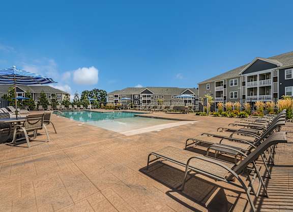 A pool surrounded by chairs and umbrellas with apartment buildings in the background.