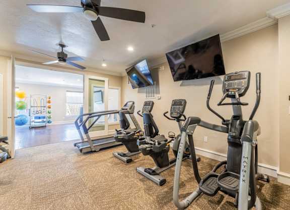 Cardio machines including an elliptical, cycling machines, and a treadmill facing a wall-mounted tv in the fitness center at Encore at Buckingham apartments in Richardson, TX