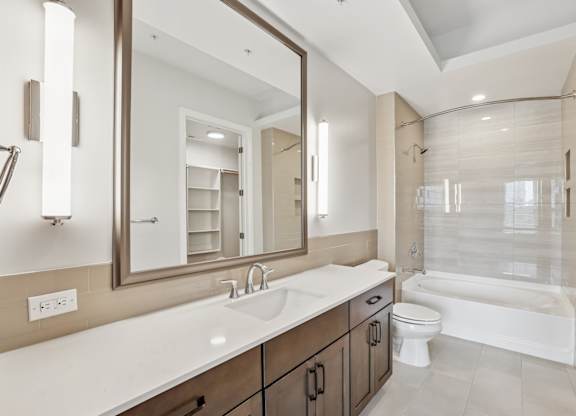 A modern bathroom from the C1 floorplan with a large mirror, white counter with wooden cabinets, sink, and bathtub at Alastair at Aria Village Apartments in Sandy Springs, GA