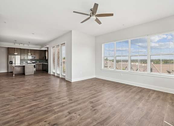 A living room from the C1 floorplan with a ceiling fan, large window, wood floors, glass doors leading to a balcony, and an open layout connecting to the kitchen at Alastair at Aria Village Apartments in Sandy Springs, GA