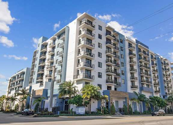 A large white and blue apartment building with balconies and palm trees in front at Alameda West Apartments in Miami, FL
