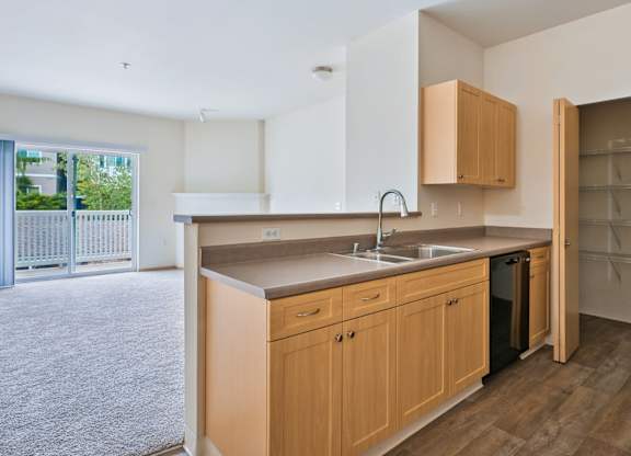 Soho kitchen with wooden cabinets, a pantry closet, and the living room in the background at Abbey Rowe Apartments in Olympia, WA