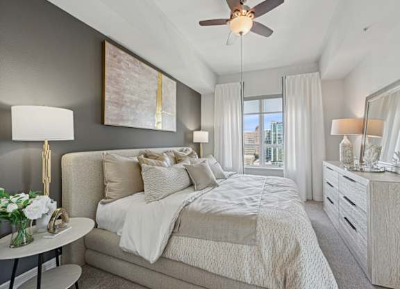 Model bedroom with a large bed, a ceiling fan, and a view of the city at Dominion Post Oak apartments in Houston, TX
