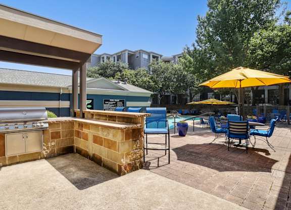 A grilling area is on a patio with blue chairs and yellow umbrellas with the pool in the background at Encore at Buckingham Apartments in Richardson, TX