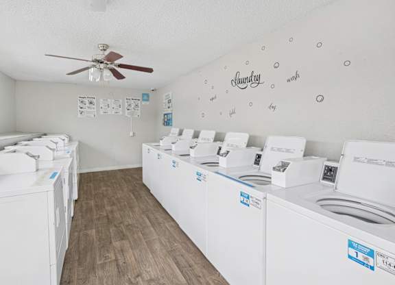 A room with a white ceiling and floor with a wall of washing machines on the right and a fan on the ceiling.