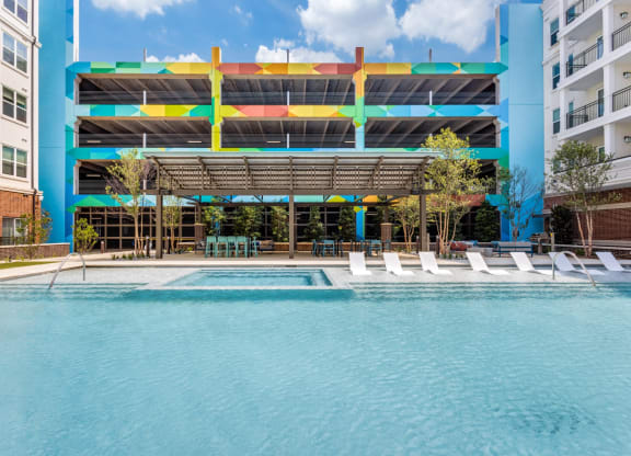 Resort-style swimming pool with in-pool white lounge chairs and parking garage in background at Era Apartments in Denton, TX