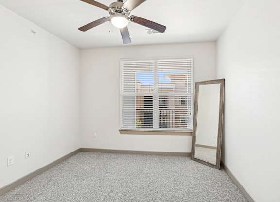 A bedroom with a ceiling fan, mirror, and a window with blinds at GreenVue Apartments in Richardson, TX