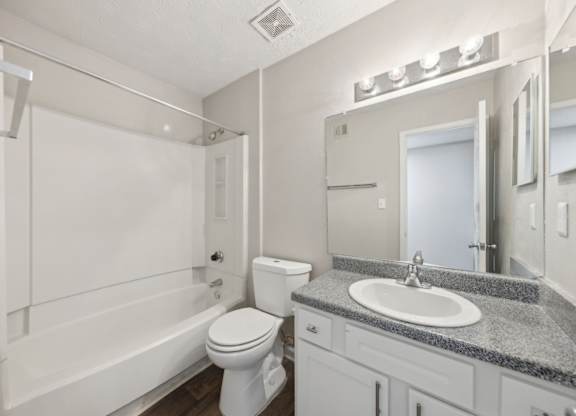 A bathroom with a white bathtub and shower, toilet, sink, and a mirror with lights at Gwinnett Square Apartments in Duluth, GA
