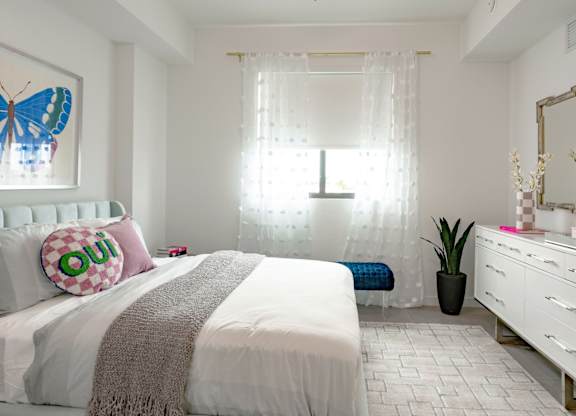 Side view of 1st bedroom in our 2-bed model with a white bed, a white dresser, and big window showering room with sunlight at Hollywood Heights on the Boulevard Luxury Apartments in Hollywood, FL