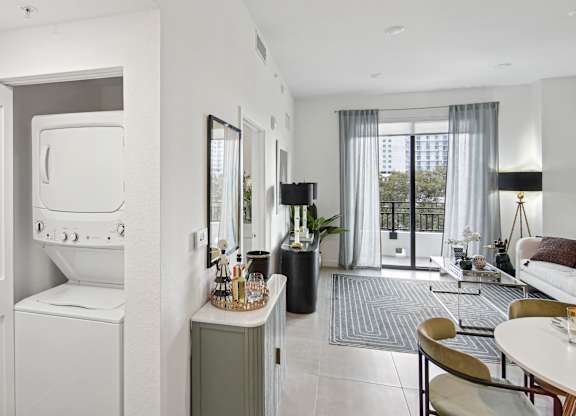 A modern living room with a window with open curtains, a dining table and chairs, and a washer and dryer  A modern kitchen with a dining table and chairs, fridge with a ice maker, stovetop, and microwave  Living room and modern kitchen in our 2-bedroom model with a dining table and chairs  Balcony view of Hollywood Boulevard at Hollywood Heights on the Boulevard Luxury Apartments in Hollywood, FL
