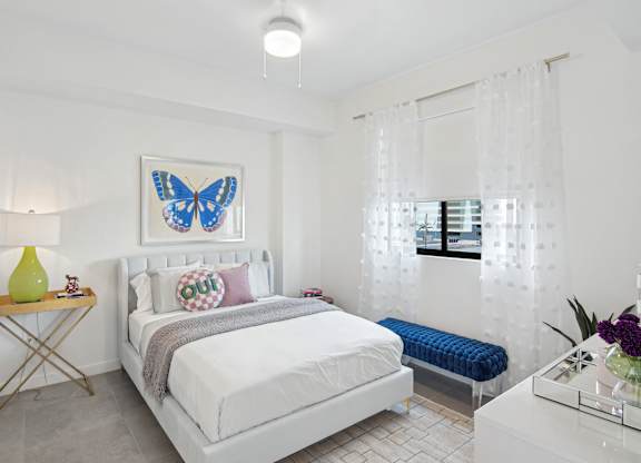 A bedroom with a bed, a blue bench, and a blue butterfly picture on the wall above the bed  Second bedroom with a bed, nightstand in our 2-bedroom model at Hollywood Heights on the Boulevard Luxury Apartments in Hollywood, FL