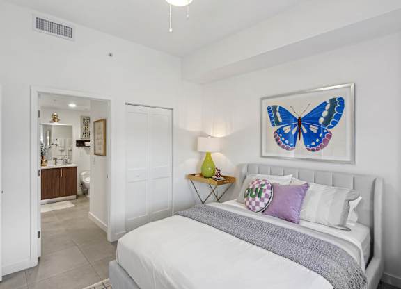 A bedroom with a bed, a blue bench, and a blue butterfly picture on the wall above the bed  Second bedroom with a bed, nightstand in our 2-bedroom model at Hollywood Heights on the Boulevard Luxury Apartments in Hollywood, FL