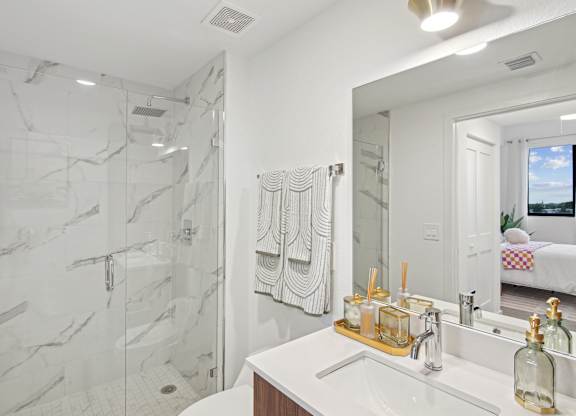 A bathroom with a marble shower and white countertop  A bedroom with a bed, a blue bench, and a blue butterfly picture on the wall above the bed  Second bedroom with a bed, nightstand in our 2-bedroom model at Hollywood Heights on the Boulevard Luxury Apartments in Hollywood, FL