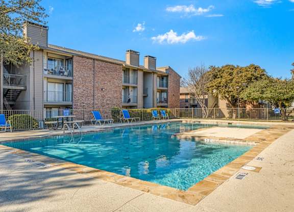 A swimming pool in front of a brick building with blue chairs around it.