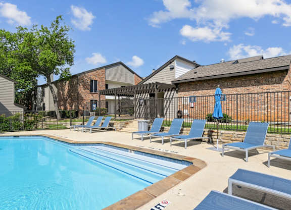 A swimming pool surrounded by lounge chairs and a fence at The Oaks of Denton Apartments in Denton, TX