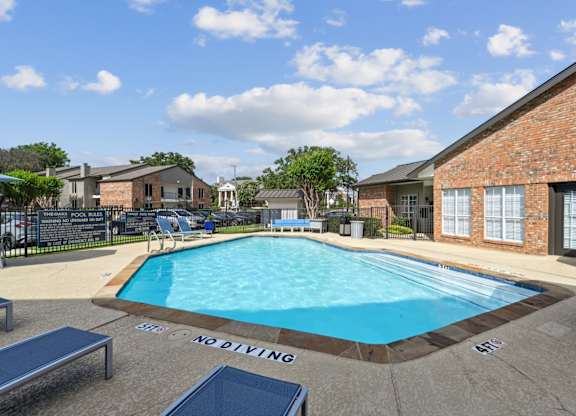 A pool with blue lounge chairs and apartment buildings in the background at The Oaks of Denton Apartments in Denton, TX