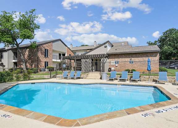 A swimming pool surrounded by chairs and a fence with the leasing office in the background at The Oaks of Denton Apartments in Denton, TX