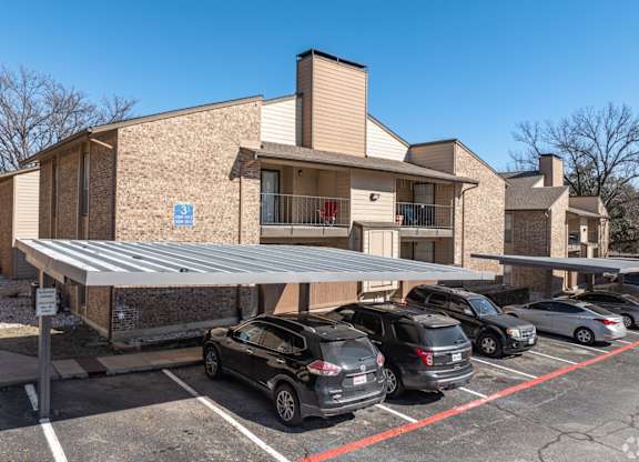 A covered parking area with cars and an apartment building in the background at Preston Oaks Apartments in Dallas, TX.