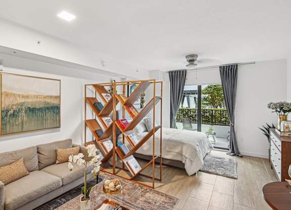 Studio Sixty2 floor plan living area with a couch, a painting, a bookshelf, bed, and a sliding door with curtains leading to balcony at Twenty2 West luxury apartments in Miami, FL