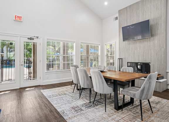 A dining room with a wooden table and chairs, a television on the wall, and a door leading outside.