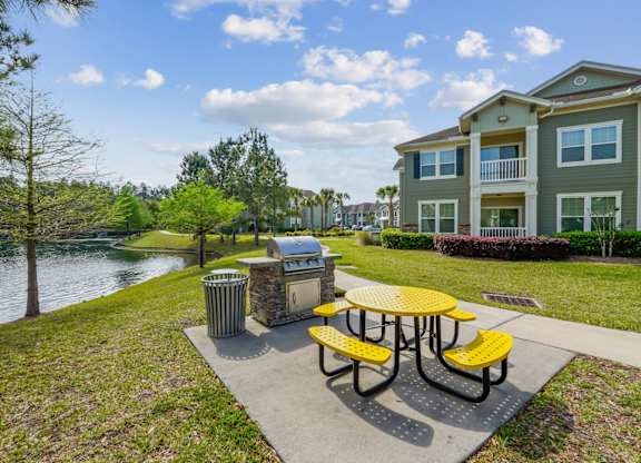 A yellow picnic table is in the foreground of a park with a lake and apartment building in the background.