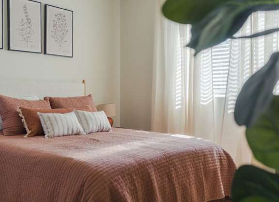 A bed with a pink comforter and two pillows is in the foreground with a white wall and two framed pictures above it.