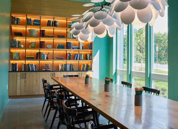 A long wooden table is surrounded by chairs in a room with a green wall and a bookshelf.