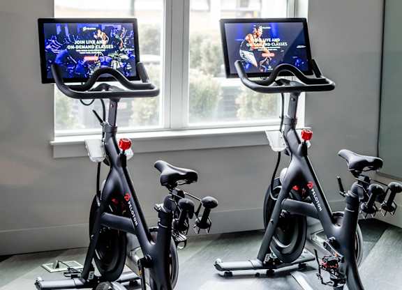Two stationary bikes in a room with screens on the handlebars.