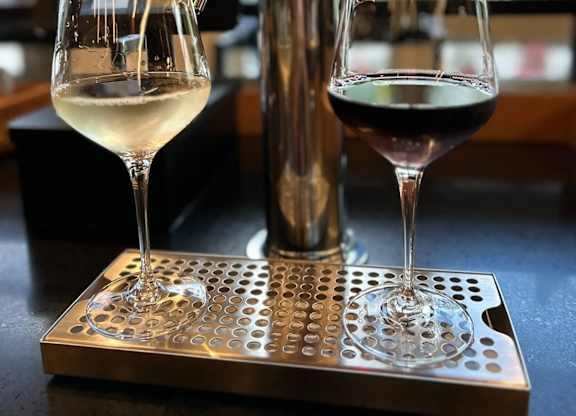 Two wine glasses, one with white wine and the other with red wine, are placed on a metal tray next to a beer tap.