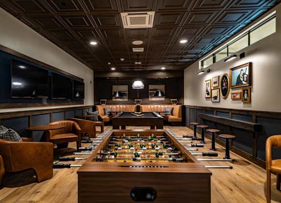 A foosball table is in the middle of a room with leather chairs and a bar area.