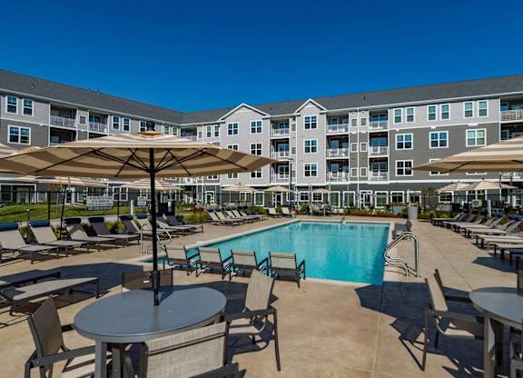 A large outdoor pool area with lounge chairs and umbrellas in front of a multi-story apartment building.