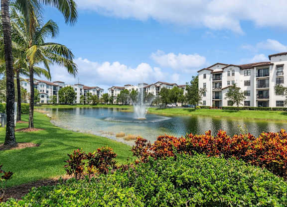 A beautiful view of a resort with a fountain in the middle of a lake surrounded by trees and buildings.