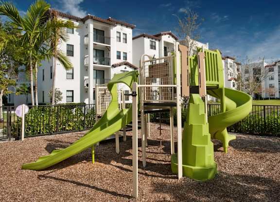 A playground with a green slide in front of apartment buildings.