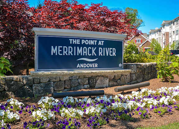 A sign that says The Point at Merrimack River Andover stands in front of flowers.