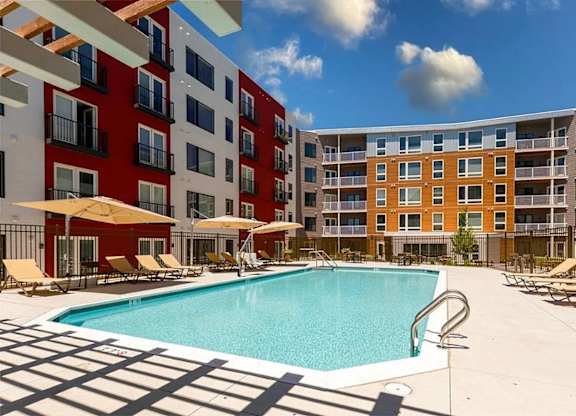 A swimming pool surrounded by sun loungers and umbrellas in front of a multi-story apartment building.