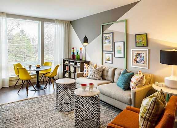 A living room with a grey couch, a black and white ottoman, a yellow chair, a black table, a bookshelf, and a window with white curtains.