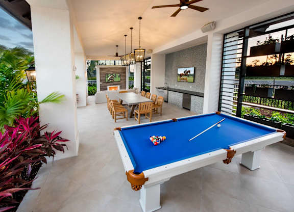 A pool table with a blue cloth and a white cue is in the center of a room with a ceiling fan and a television.