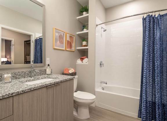 A bathroom with a white toilet, a sink with a marble countertop, a shower with a blue curtain, and a mirror above the sink.