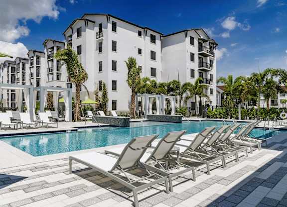 A row of lounge chairs are set up by a pool in front of a white apartment building.