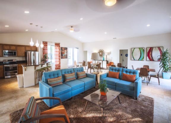 Apartments for Rent in Goleta, CA - Pacific Oaks - Community Room with Couches, Armchair, Coffee Table, Side Table with Lamp, Dining Tables and Chairs, Kitchen with Stainless Steel Appliances, Breakfast Bar, Bar Stools, and Pendant Lights.