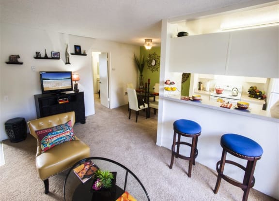 Goleta, CA, Apartments Near UCSB - Pacific Oaks - Living Room with Open Floor Plan, Carpeting, Chair, Glass-Top Coffee Table, TV, Breakfast Bar, Barstools, and View of Kitchen and Dining Area.