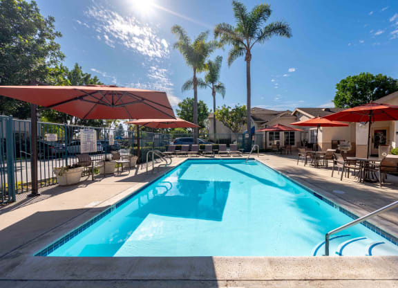 Ventura, CA, Apartments Near The Collection Oxnard - Cypress Meadows - Swimming Pool With Red Umbrellas, Lounge Chairs, Patio Tables, And Palm Trees Beside A Clubhouse.