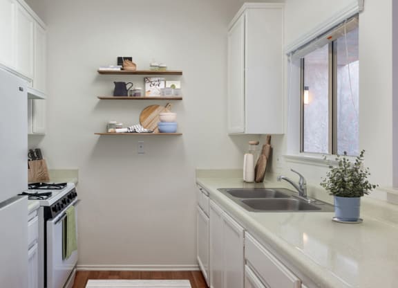 One-Bedroom Apartments in Ventura, CA - Cypress Meadows - Kitchen Features White Cabinets, A Double Sink, Open Wall Shelves, And White Appliances.