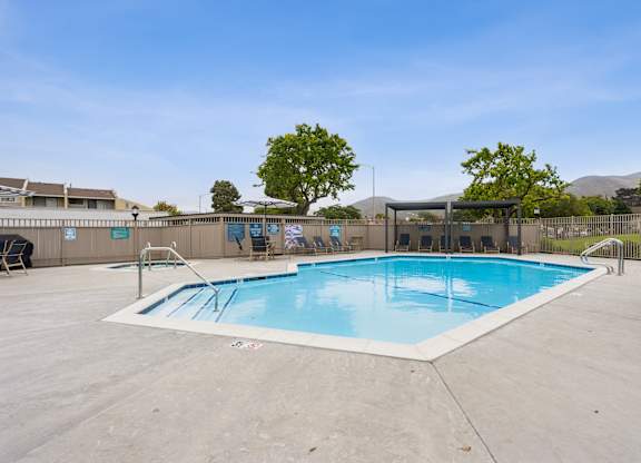 Lompoc, CA, Apartments - Oceanwood Apartments - A Large Outdoor Swimming Pool Features A Wide Concrete Deck, Metal Handrails, Lounge Seating Under Shade Structures, And A Fenced Perimeter.