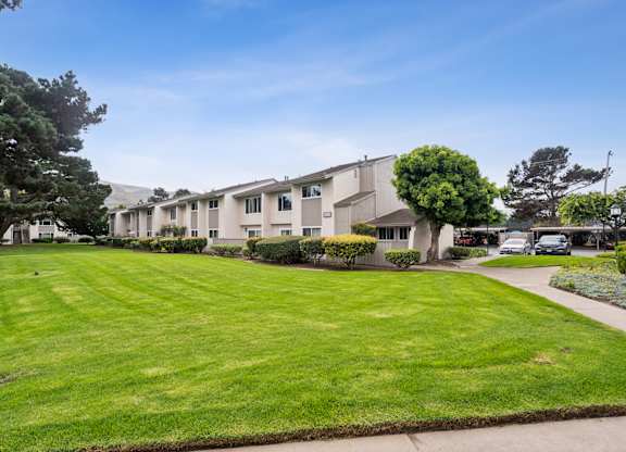 Lompoc, CA, Apartments Near Walmart - Oceanwood Apartments - Community Courtyard With Trimmed Shrubs, Mature Trees, And A Curved Walkway Leading Toward Parking.