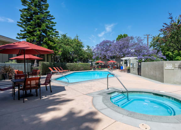 Apartments in Goleta, CA for Rent - Encina Meadows - Pool with Hot Tub, Poolside Lounge Seating, Tables and Chairs with Umbrellas, and Surrounding Hedges and Trees.